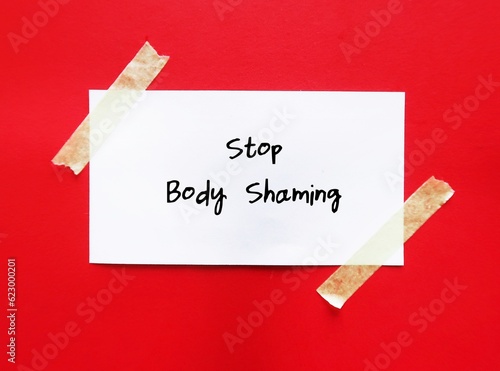 Note stick on red background with handwritten text STOP BODY SHAMING , no more humiliation about another body shape , size or criticizing appearance through negative judgment