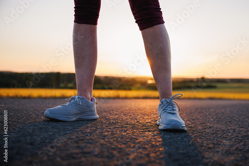 Woman with running shoe standing on road at sunset. Sport training