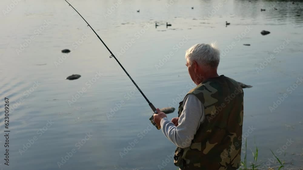Old man fishing. Senior gray haired fisherman throws a spinning from shoreside at sunset, twisting a coil. Positive elderly male angling at lake, rotating reel rod. Retired fisher enjoying his weekend Old man fishing. Senior gray haired fisherman throws a spinning from shoreside at sunset, twisting a coil. Positive elderly male angling at lake, rotating reel rod. Retired fisher enjoying his weekend