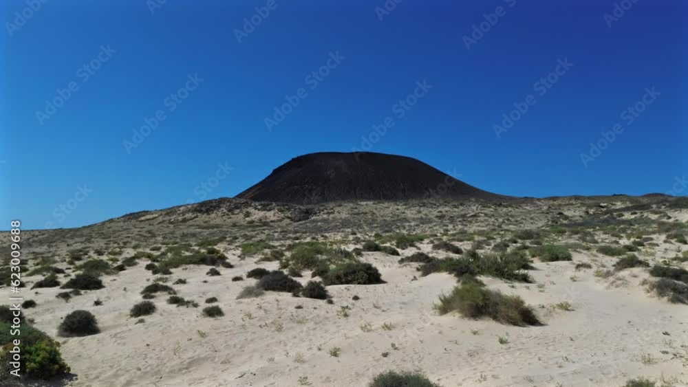 A drone video capturing a sand landscape with low vegetation, flying at ground level and gradually revealing a volcano in the background under a clear blue sky A drone video capturing a sand landscape with low vegetation, flying at ground level and gradually revealing a volcano in the background under a clear blue sky