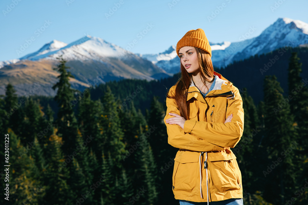 Woman hiker standing on the mountain looking at nature happiness with a view of snowy mountains and trees in yellow raincoat and cap travel autumn and hiking in the mountains at sunset freedom