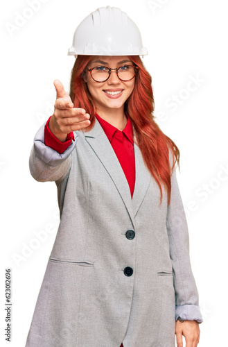 Young redhead woman wearing architect hardhat smiling friendly offering handshake as greeting and welcoming. successful business.