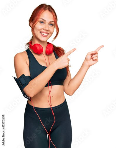 Young redhead woman wearing gym clothes and using headphones smiling and looking at the camera pointing with two hands and fingers to the side.