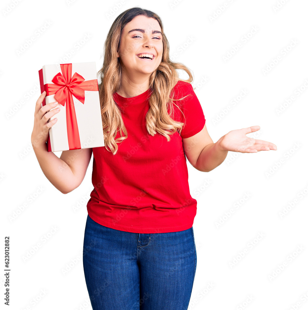 Young caucasian woman holding gift celebrating victory with happy smile and winner expression with raised hands Young caucasian woman holding gift celebrating victory with happy smile and winner expression with raised hands