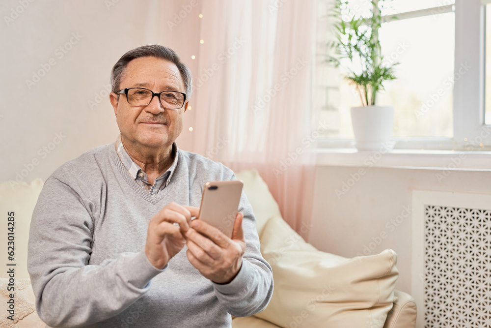 People And Technology. Portrait of smiling mature man using mobile phone, watching video or reading sms message, sitting on the couch in living room at home. Elderly man uses modern technology People And Technology. Portrait of smiling mature man using mobile phone, watching video or reading sms message, sitting on the couch in living room at home. Elderly man uses modern technology