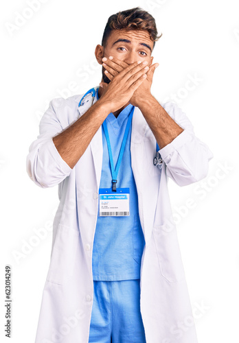 Young hispanic man wearing doctor uniform and stethoscope shocked covering mouth with hands for mistake. secret concept.