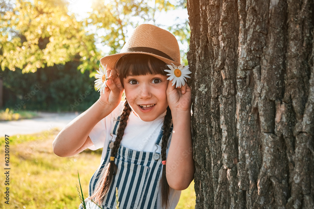 Cute little blonde girl in a cotton dress and straw hat walks in a field of daisies collects them in the basket