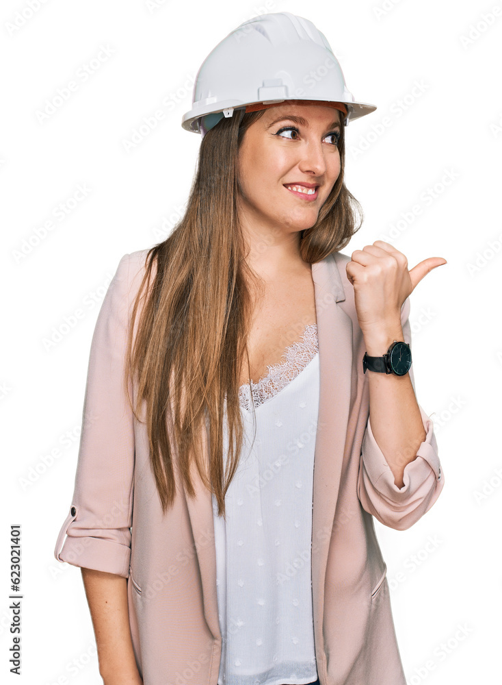 Young blonde woman wearing architect hardhat smiling with happy face looking and pointing to the side with thumb up. Young blonde woman wearing architect hardhat smiling with happy face looking and pointing to the side with thumb up.