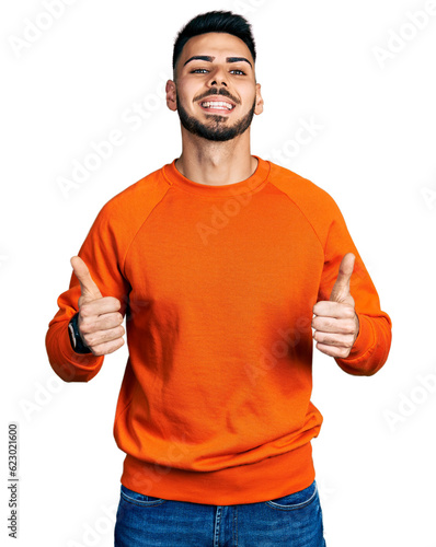 Young hispanic man with beard wearing casual orange sweater success sign doing positive gesture with hand, thumbs up smiling and happy. cheerful expression and winner gesture. © Krakenimages.com