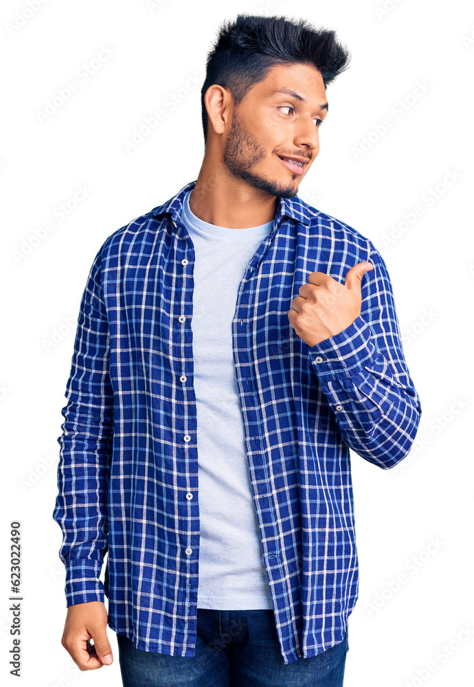 Handsome latin american young man wearing casual shirt smiling with happy face looking and pointing to the side with thumb up. Handsome latin american young man wearing casual shirt smiling with happy face looking and pointing to the side with thumb up.
