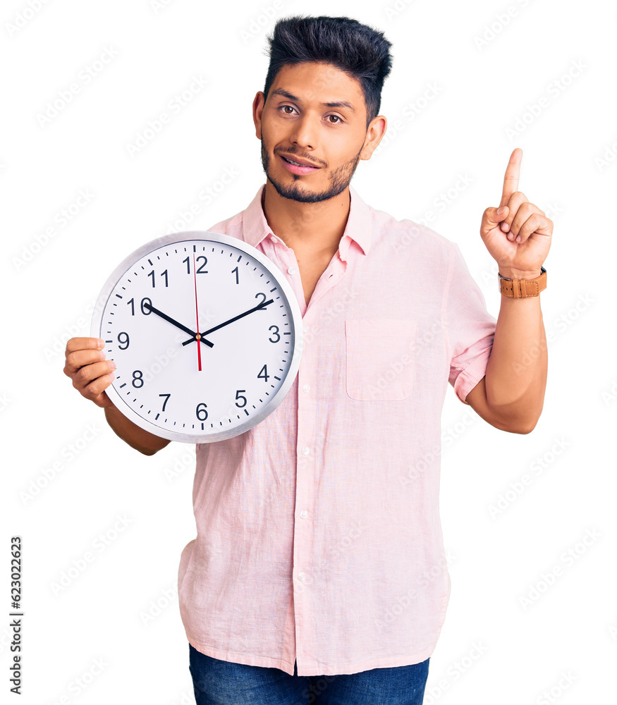 Handsome latin american young man holding big clock surprised with an idea or question pointing finger with happy face, number one Handsome latin american young man holding big clock surprised with an idea or question pointing finger with happy face, number one