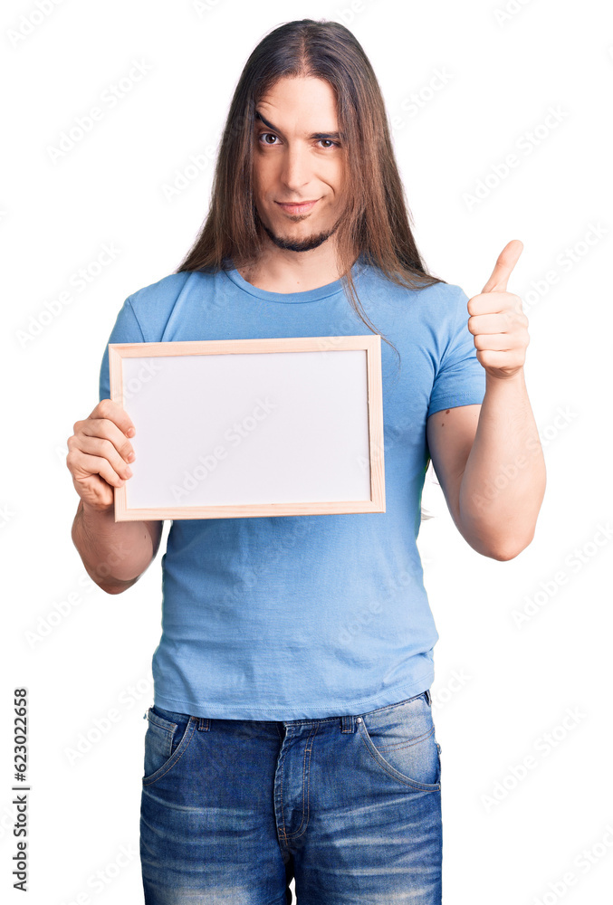 Young adult man with long hair holding empty white chalkboard smiling happy and positive, thumb up doing excellent and approval sign Young adult man with long hair holding empty white chalkboard smiling happy and positive, thumb up doing excellent and approval sign