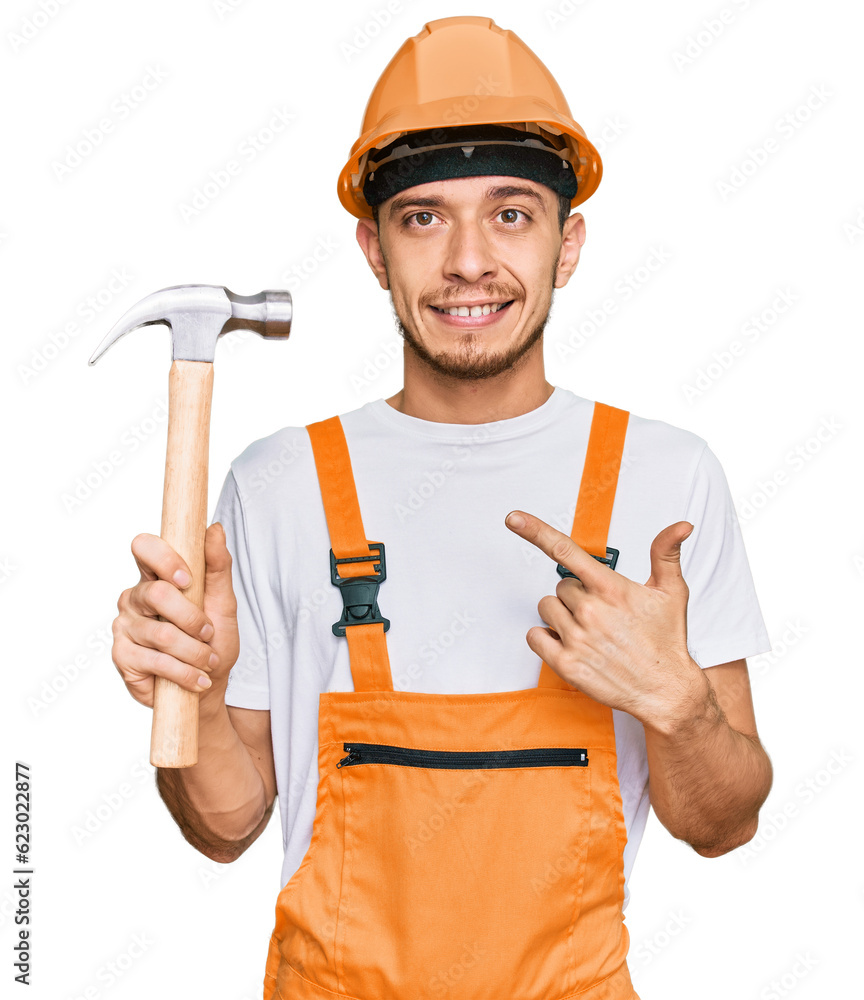 Hispanic young man wearing hardhat holding hammer smiling happy pointing with hand and finger