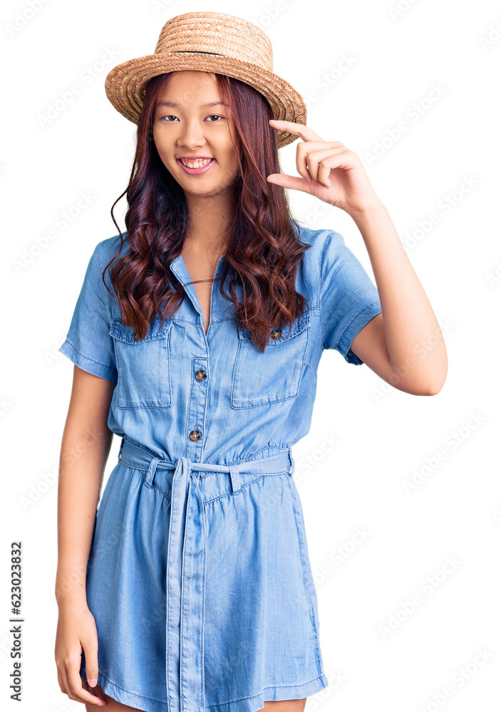 Young beautiful chinese girl wearing summer hat smiling and confident gesturing with hand doing small size sign with fingers looking and the camera. measure concept. Young beautiful chinese girl wearing summer hat smiling and confident gesturing with hand doing small size sign with fingers looking and the camera. measure concept.