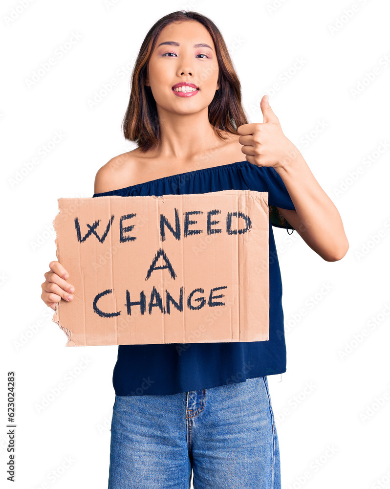 Young beautiful chinese girl holding we need a change banner smiling happy and positive, thumb up doing excellent and approval sign Young beautiful chinese girl holding we need a change banner smiling happy and positive, thumb up doing excellent and approval sign