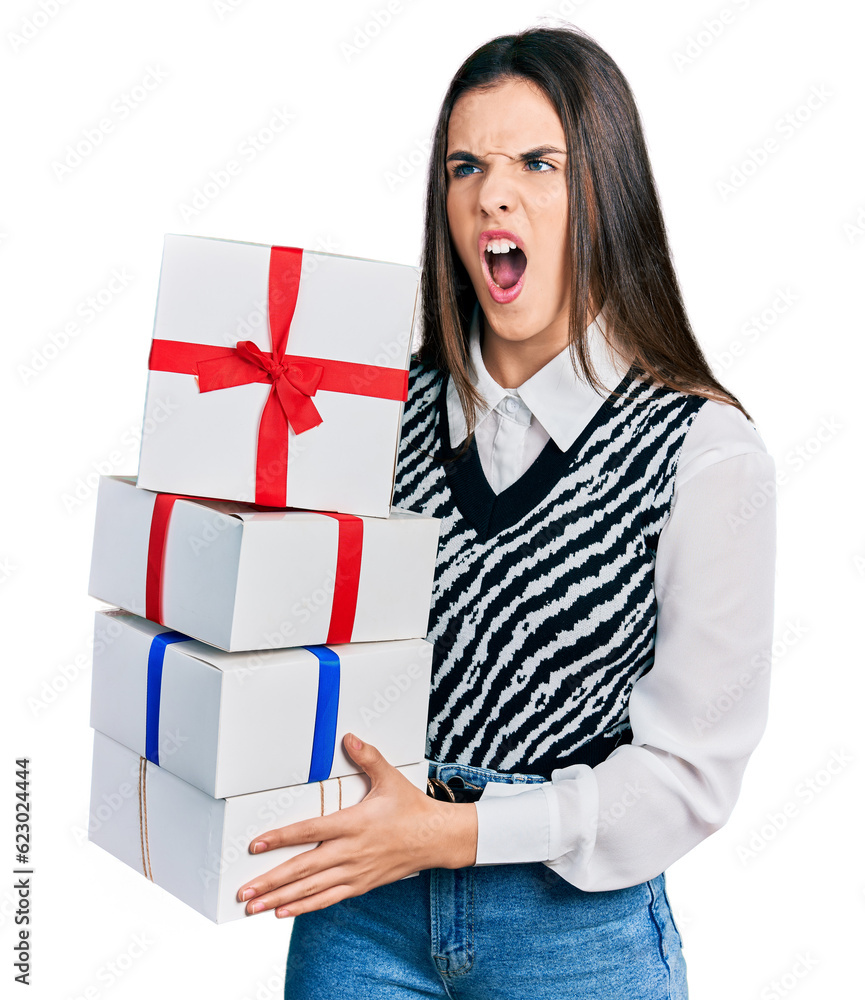 Young brunette teenager holding gifts angry and mad screaming frustrated and furious, shouting with anger. rage and aggressive concept. Young brunette teenager holding gifts angry and mad screaming frustrated and furious, shouting with anger. rage and aggressive concept.