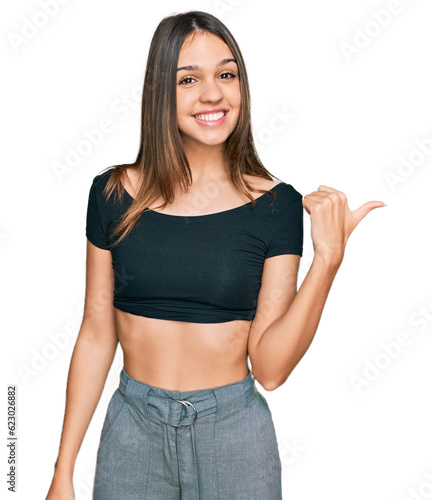 Young brunette woman wearing casual clothes smiling with happy face looking and pointing to the side with thumb up.
