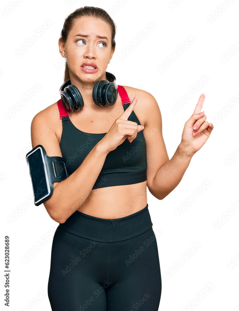 Beautiful blonde woman wearing gym clothes and using headphones pointing aside worried and nervous with both hands, concerned and surprised expression Beautiful blonde woman wearing gym clothes and using headphones pointing aside worried and nervous with both hands, concerned and surprised expression
