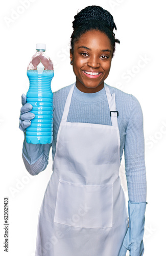 Young african american woman wearing apron holding detergent bottle looking positive and happy standing and smiling with a confident smile showing teeth