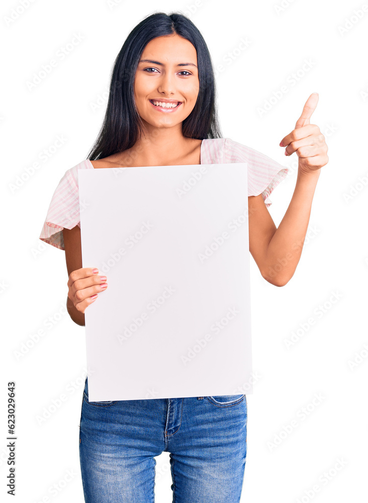 Young beautiful latin girl holding blank empty banner smiling happy and positive, thumb up doing excellent and approval sign Young beautiful latin girl holding blank empty banner smiling happy and positive, thumb up doing excellent and approval sign