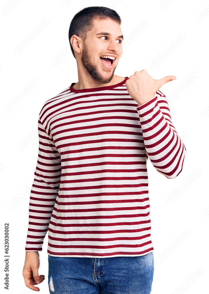 Young handsome man wearing striped sweater smiling with happy face looking and pointing to the side with thumb up. Young handsome man wearing striped sweater smiling with happy face looking and pointing to the side with thumb up.