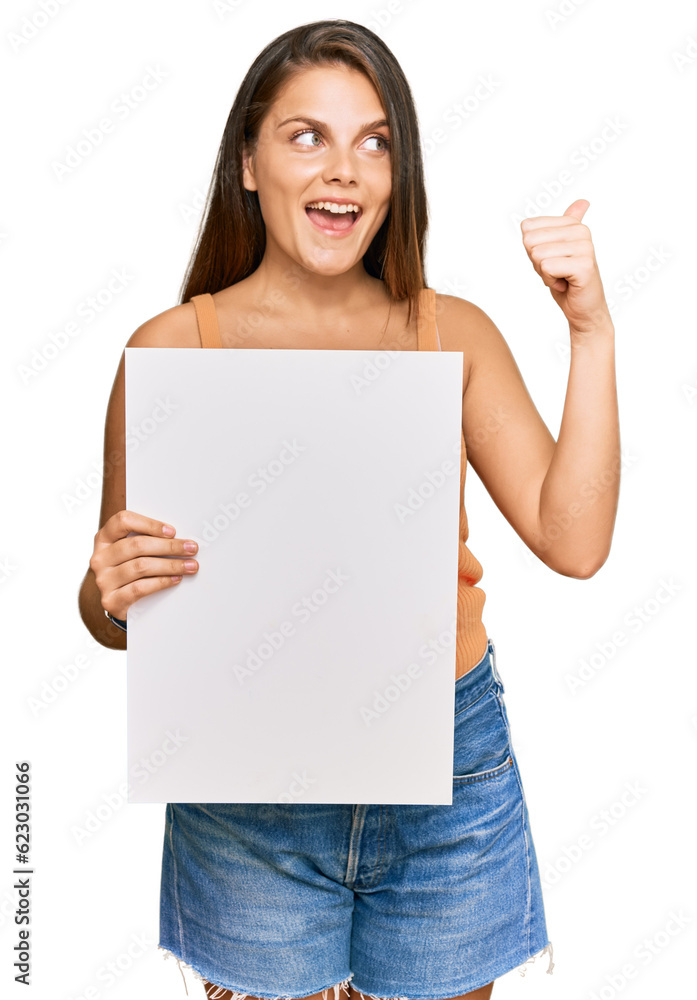 Young caucasian woman holding blank empty banner pointing thumb up to the side smiling happy with open mouth Young caucasian woman holding blank empty banner pointing thumb up to the side smiling happy with open mouth