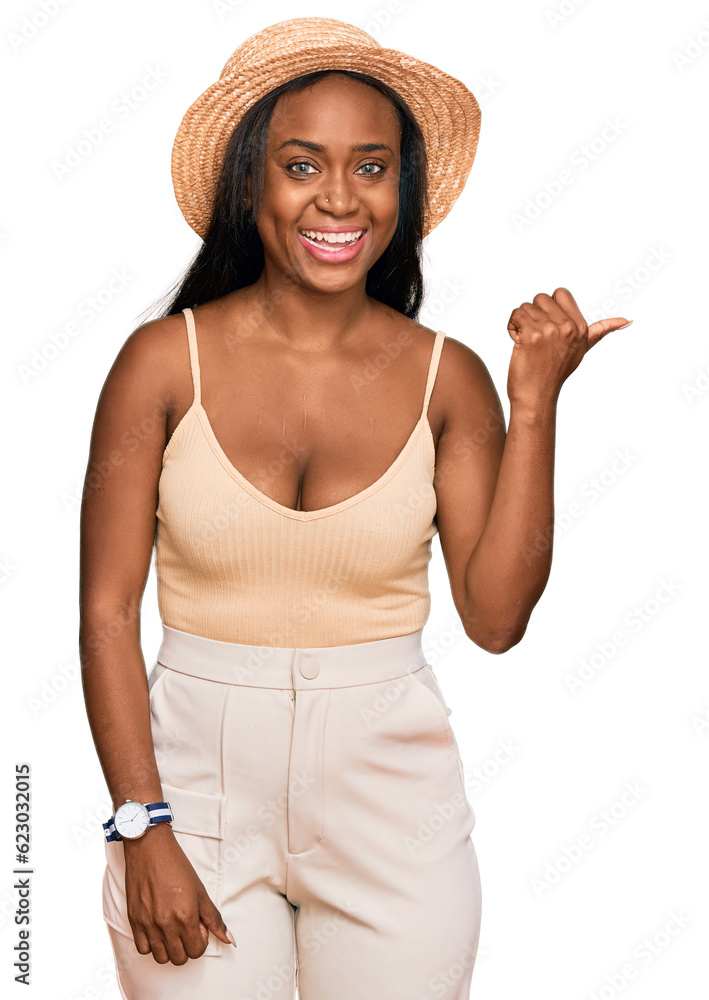 Young black woman wearing summer hat smiling with happy face looking and pointing to the side with thumb up. Young black woman wearing summer hat smiling with happy face looking and pointing to the side with thumb up.