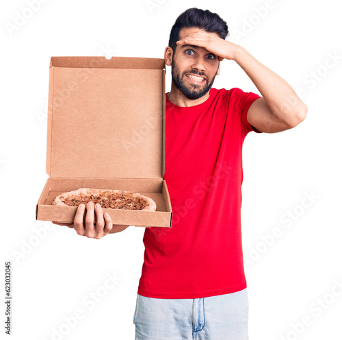 Young handsome man with beard holding delivery cardoboard with italian pizza stressed and frustrated with hand on head, surprised and angry face photo