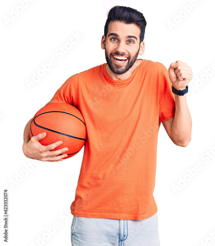 Young handsome man with beard hoilding basketball ball screaming proud, celebrating victory and success very excited with raised arms © Krakenimages.com