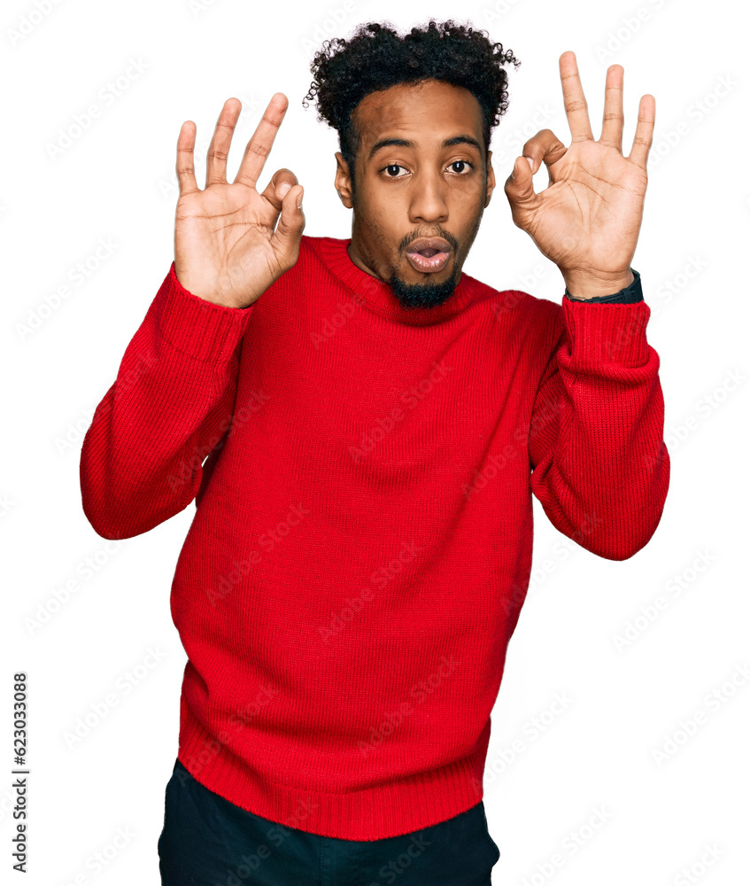 Young african american man with beard wearing casual winter sweater looking surprised and shocked doing ok approval symbol with fingers. crazy expression Young african american man with beard wearing casual winter sweater looking surprised and shocked doing ok approval symbol with fingers. crazy expression