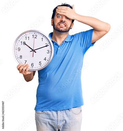 Young hispanic man holding big clock stressed and frustrated with hand on head, surprised and angry face