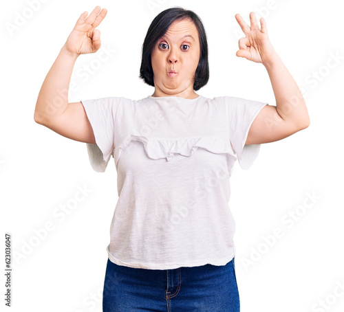 Brunette woman with down syndrome wearing casual white tshirt looking surprised and shocked doing ok approval symbol with fingers. crazy expression