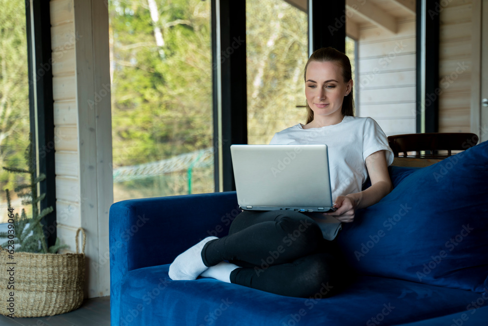 Naklejka premium A young brunette is working with a laptop, sitting on a blue sofa at home.