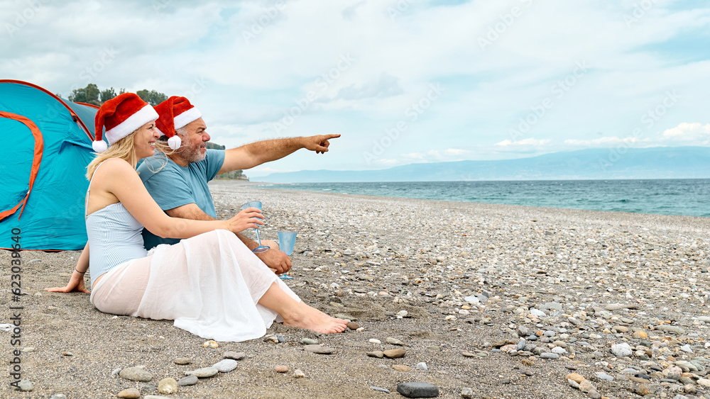 Middle aged couple wearing santa hats having christmas picnic on the beach. Bearded man and pretty blond woman drinking sparkling wine and having fun looking the sea. Middle aged couple wearing santa hats having christmas picnic on the beach. Bearded man and pretty blond woman drinking sparkling wine and having fun looking the sea.