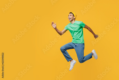 Full body side view excited young man of African American ethnicity wear casual clothes green t-shirt hat jump high look aside isolated on plain yellow background studio portrait. Lifestyle concept.
