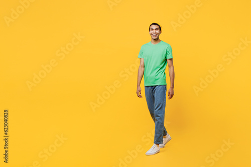 Full body smiling happy cheerful young man of African American ethnicity he wears casual clothes green t-shirt hat walking going isolated on plain yellow background studio portrait. Lifestyle concept.