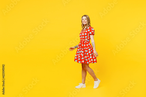 Full body side profile view young happy smiling caucasian woman she wearing red dress casual clothes walking going look camera isolated on plain yellow background studio portrait. Lifestyle concept.