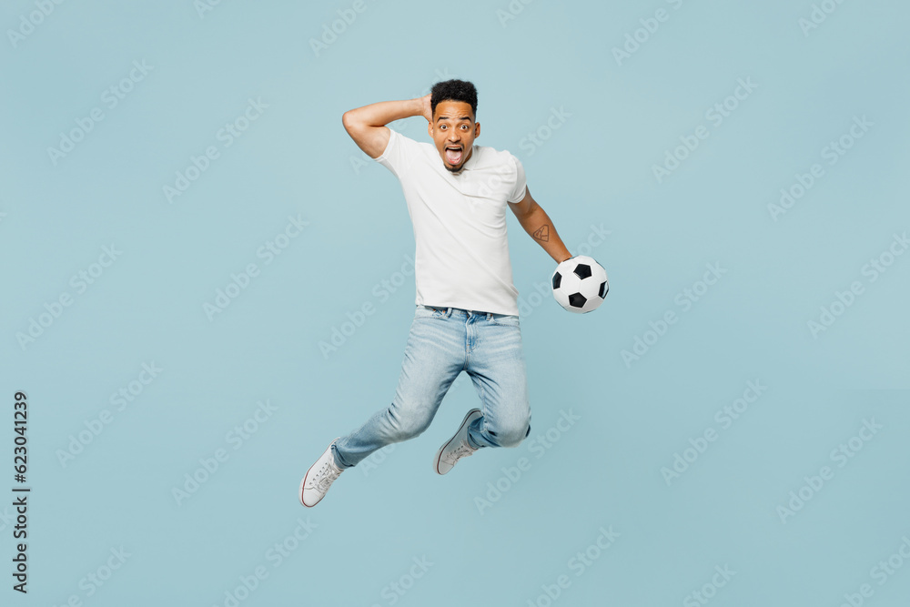 Obraz premium Full body shocked overjoyed happy fun young man fan wears t-shirt cheer up support football sport team hold soccer ball jump high watch tv live stream isolated on plain pastel blue color background.