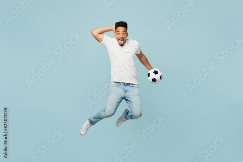 Full body shocked overjoyed happy fun young man fan wears t-shirt cheer up support football sport team hold soccer ball jump high watch tv live stream isolated on plain pastel blue color background. © ViDi Studio