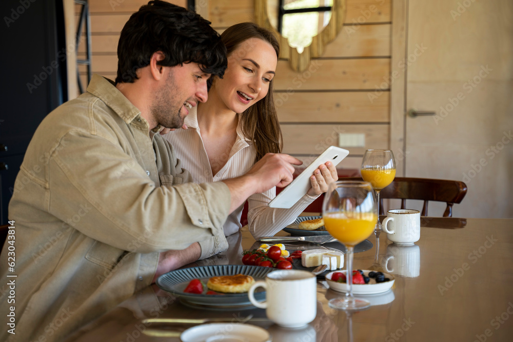 Portrait of a young man and woman looking at a digital tablet and discussing during dinner with each other, sitting at a large table with food.