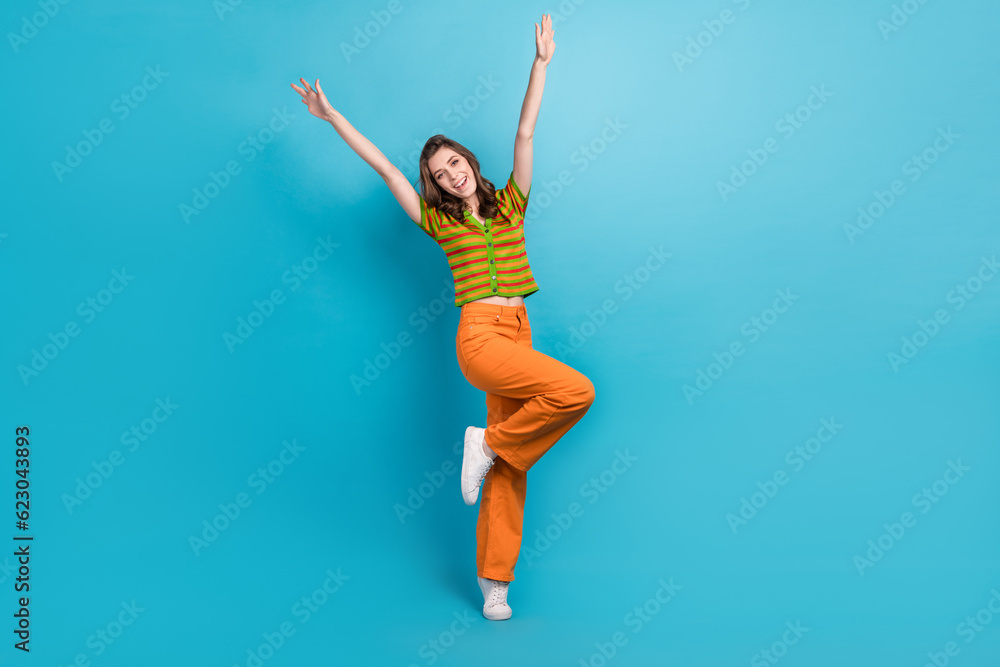 Full length photo of funny gorgeous cheerful girl wear striped shirt orange trousers raising hands up isolated on blue color background