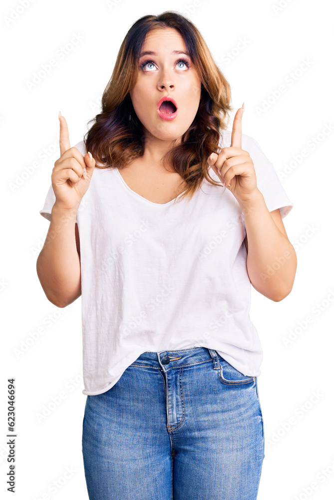 Young beautiful caucasian woman wearing casual white tshirt amazed and surprised looking up and pointing with fingers and raised arms. Young beautiful caucasian woman wearing casual white tshirt amazed and surprised looking up and pointing with fingers and raised arms.