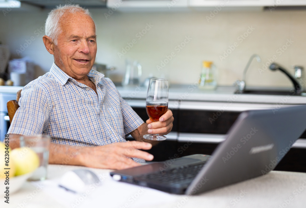 Happy elderly man with glass of wine at laptop sitting at table