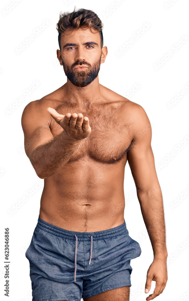 Young hispanic man wearing swimwear shirtless looking at the camera blowing a kiss with hand on air being lovely and sexy. love expression. Young hispanic man wearing swimwear shirtless looking at the camera blowing a kiss with hand on air being lovely and sexy. love expression.