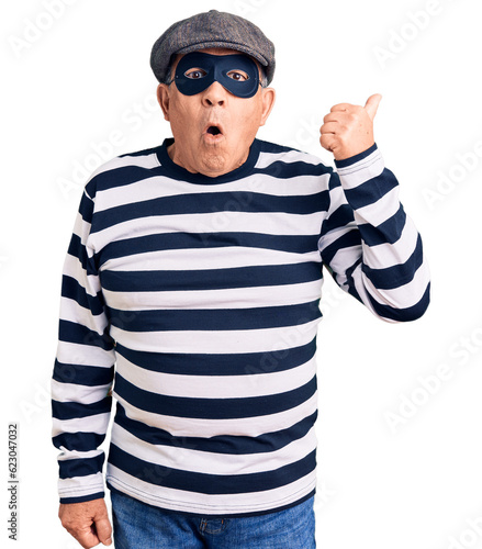 Senior handsome man wearing burglar mask and t-shirt surprised pointing with hand finger to the side, open mouth amazed expression. © Krakenimages.com