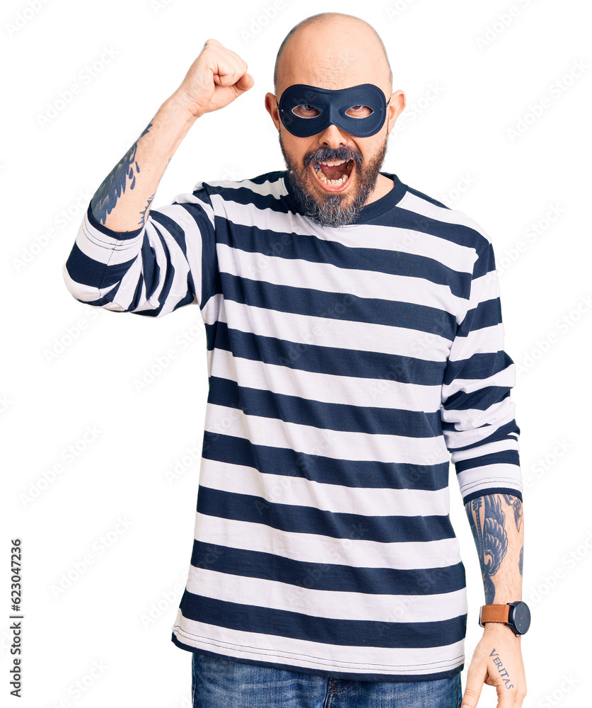 Young handsome man wearing burglar mask angry and mad raising fist frustrated and furious while shouting with anger. rage and aggressive concept. Young handsome man wearing burglar mask angry and mad raising fist frustrated and furious while shouting with anger. rage and aggressive concept.