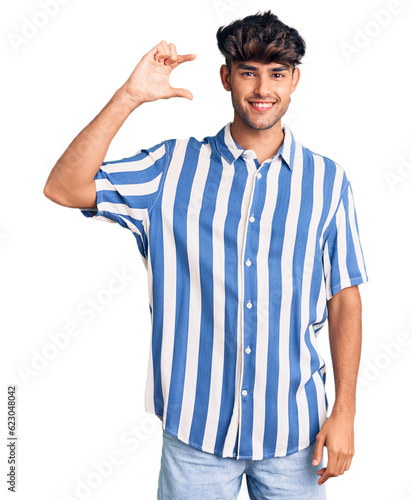 Young hispanic man wearing casual clothes smiling and confident gesturing with hand doing small size sign with fingers looking and the camera. measure concept.