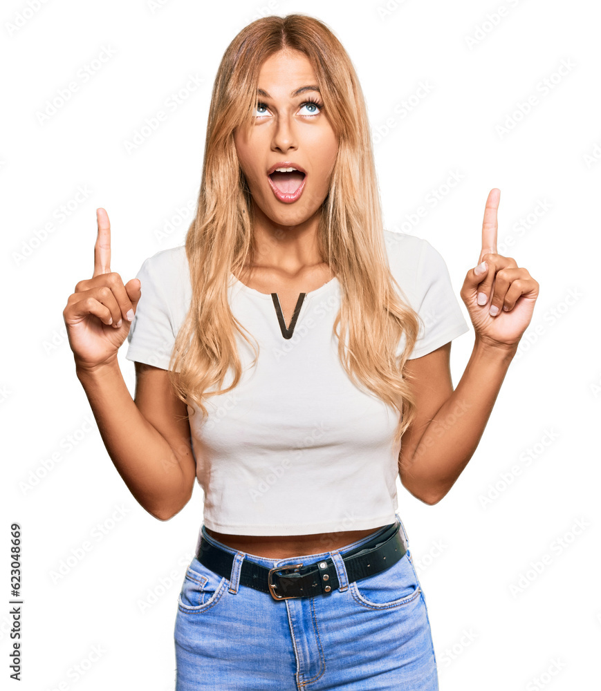 Beautiful blonde young woman wearing casual white tshirt amazed and surprised looking up and pointing with fingers and raised arms. Beautiful blonde young woman wearing casual white tshirt amazed and surprised looking up and pointing with fingers and raised arms.