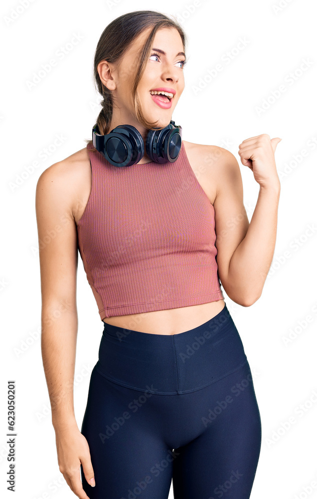 Beautiful caucasian young woman wearing gym clothes and using headphones smiling with happy face looking and pointing to the side with thumb up. Beautiful caucasian young woman wearing gym clothes and using headphones smiling with happy face looking and pointing to the side with thumb up.