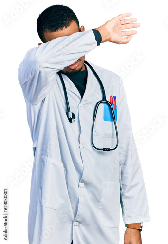 Young african american man wearing doctor uniform and stethoscope covering eyes with arm, looking serious and sad. sightless, hiding and rejection concept